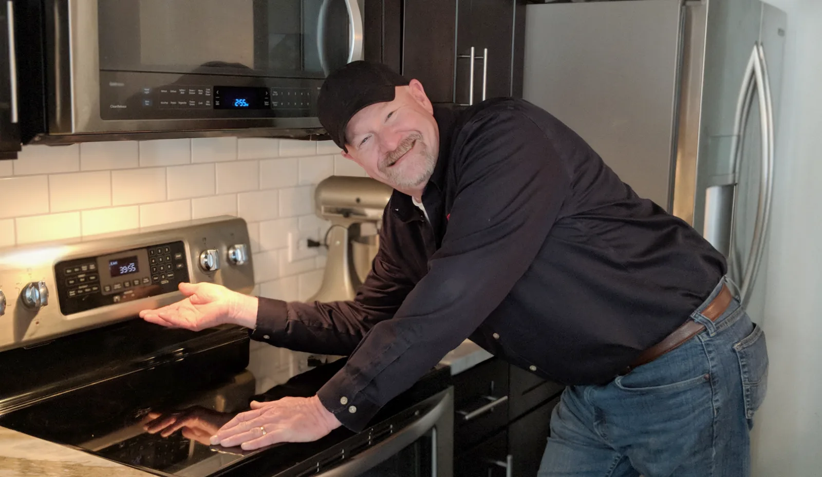 Tom Feggestad in a customer's kitchen, smiling and gesturing at a stainless glass-top range under an over-the-range microwave